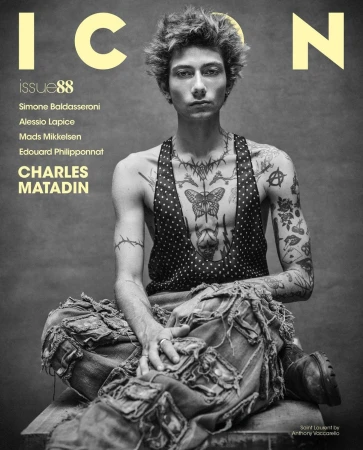 Magazine cover featuring a young adult male with tattoos, wearing a vest and pants, posed in a studio against a grey background.