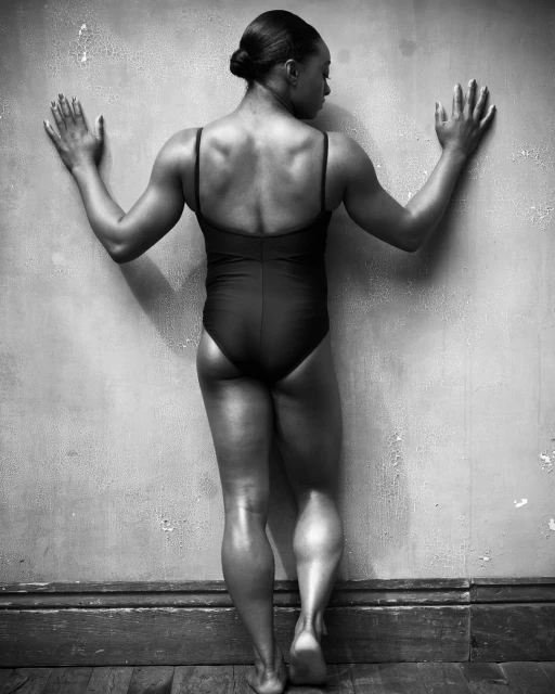 Black and white studio shot of an athletic adult female in a bodysuit, standing with arms outstretched touching a textured wall.