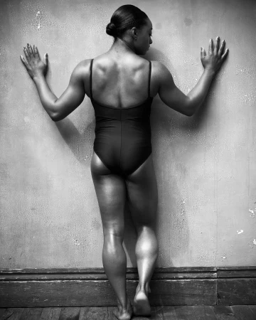 Black and white studio shot of an athletic adult female in a bodysuit, standing with arms outstretched touching a textured wall.