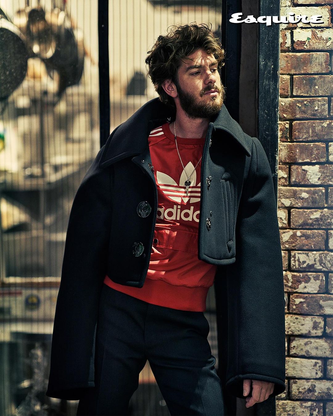 Andrew Garfield poses in a black coat and red Adidas shirt against a brick wall for Esquire magazine, showcasing a fashion-forward look.