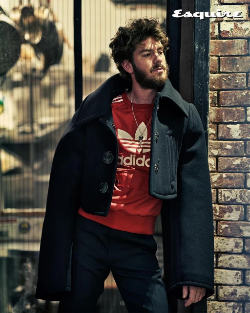 Andrew Garfield poses in a black coat and red Adidas shirt against a brick wall for Esquire magazine, showcasing a fashion-forward look.