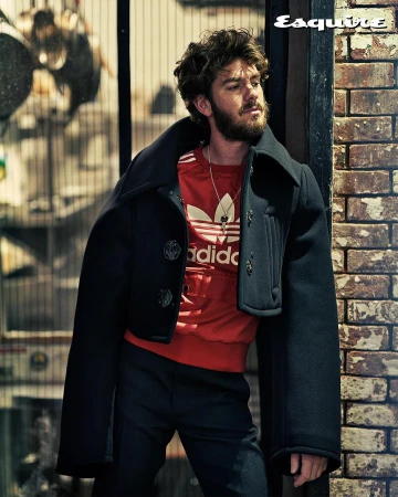 Andrew Garfield poses in a black coat and red Adidas shirt against a brick wall for Esquire magazine, showcasing a fashion-forward look.