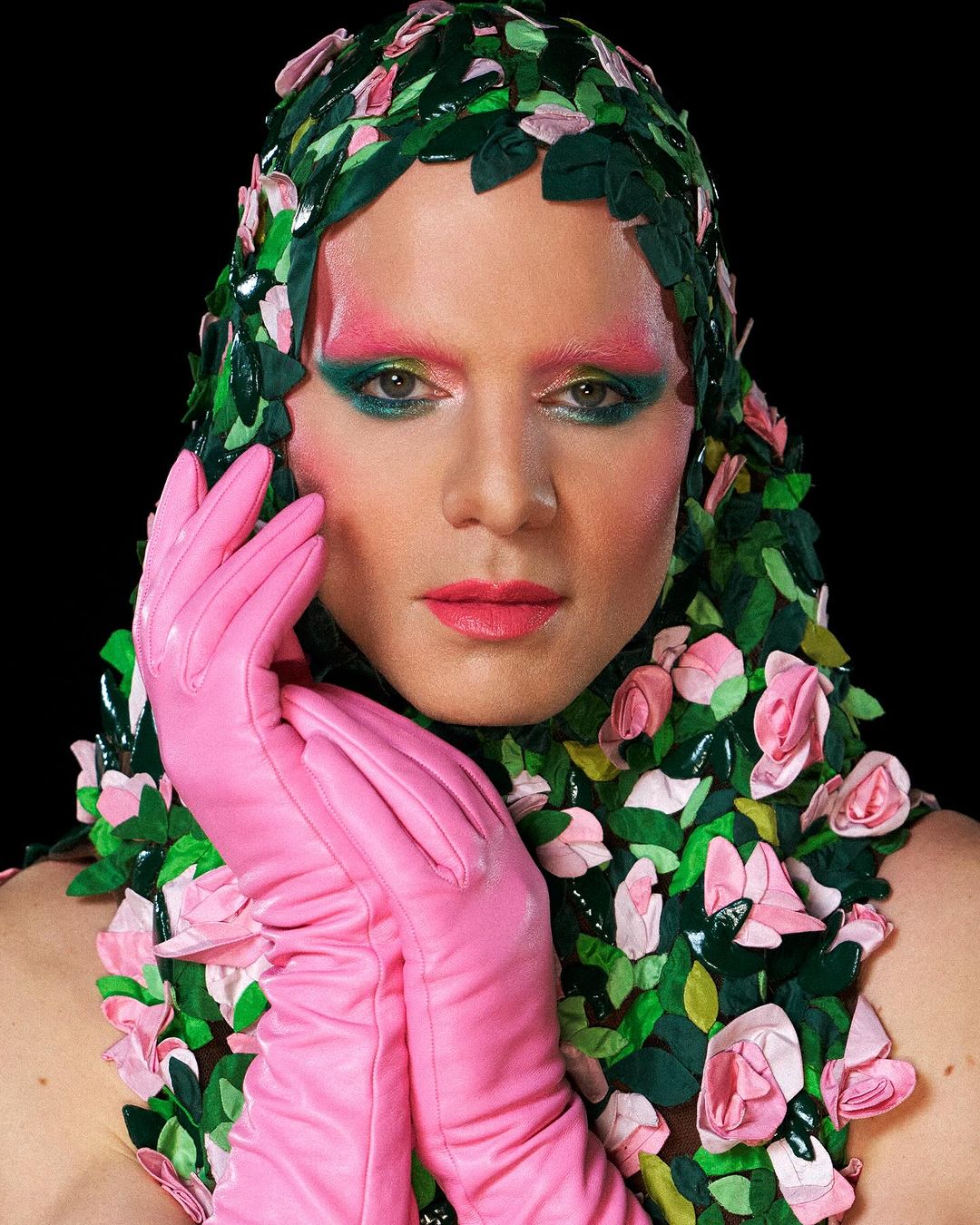 Headshot of person with theatrical makeup, pink gloves, and a headdress made of green leaves and pink roses against a black backdrop.