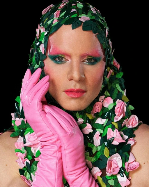 Headshot of person with theatrical makeup, pink gloves, and a headdress made of green leaves and pink roses against a black backdrop.