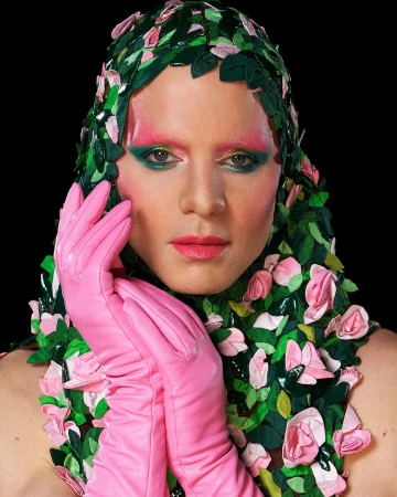 Headshot of person with theatrical makeup, pink gloves, and a headdress made of green leaves and pink roses against a black backdrop.
