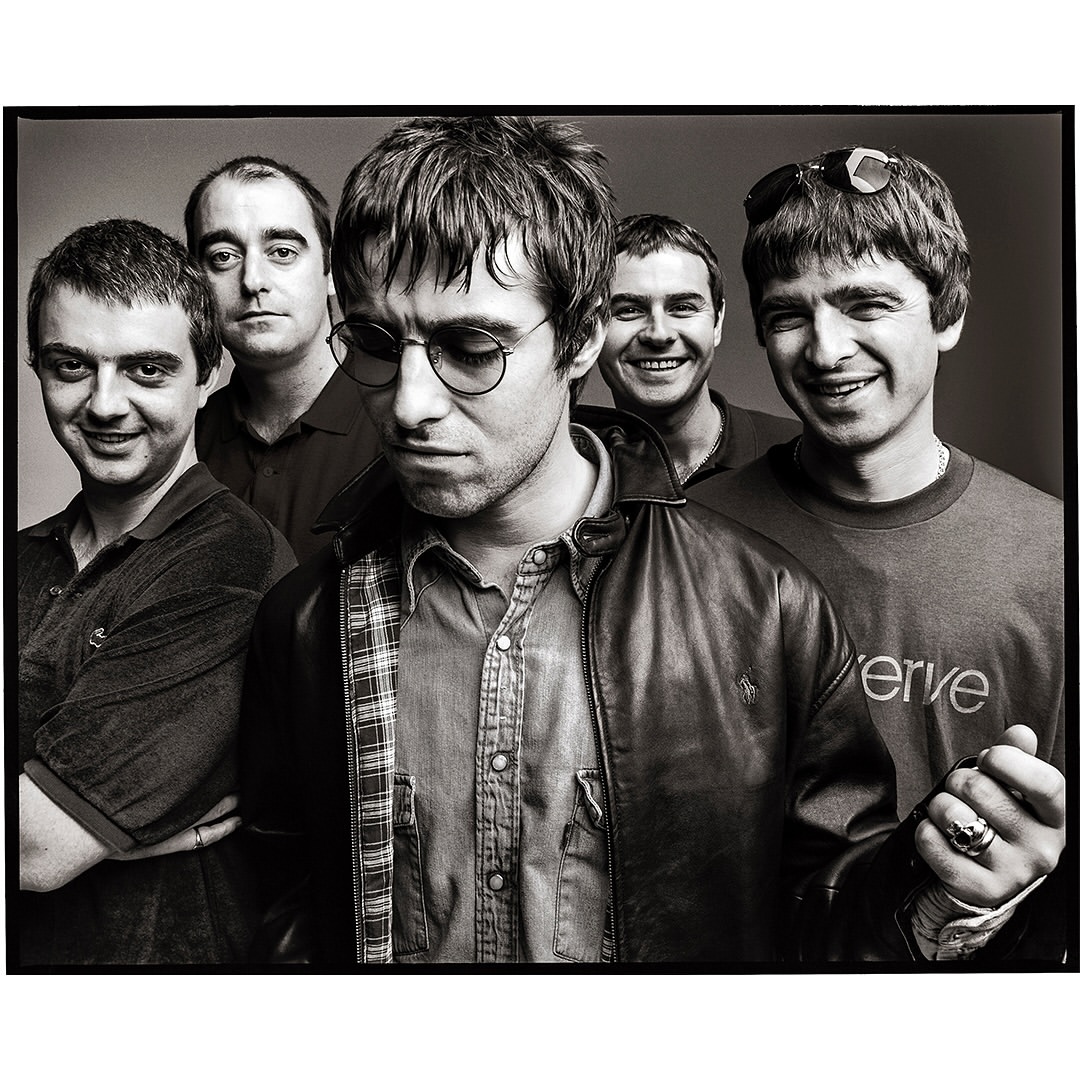 Black and white portrait of the band Oasis, featuring five members in a close-up studio shot, showcasing their individual styles and expressions.