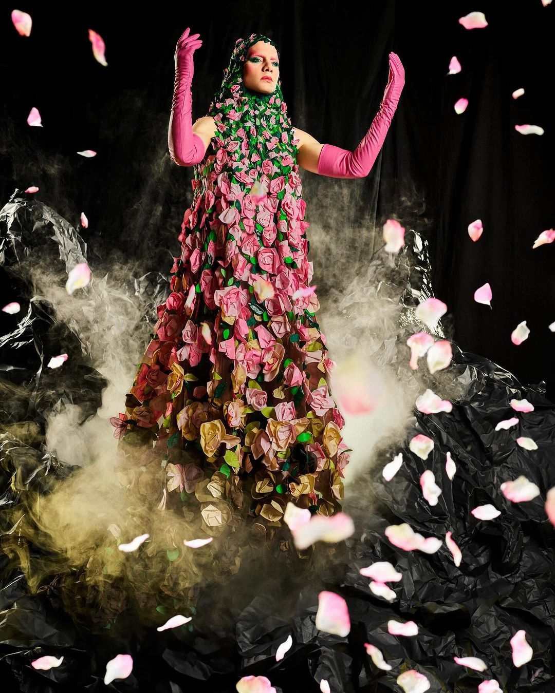 Woman in a rose-covered gown and pink gloves stands amidst fog and scattered rose petals against a dark backdrop. Dramatic fashion shot.
