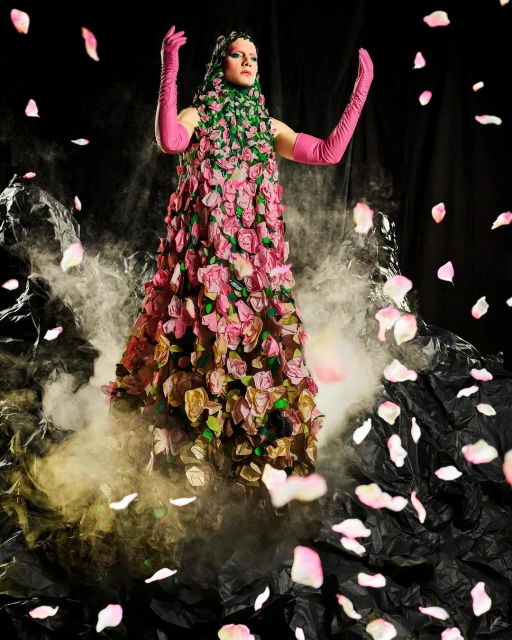 Woman in a rose-covered gown and pink gloves stands amidst fog and scattered rose petals against a dark backdrop. Dramatic fashion shot.