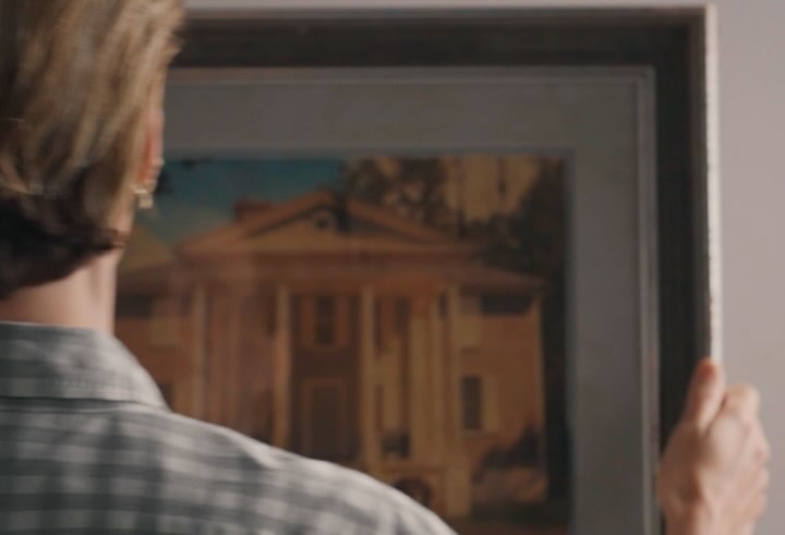 Close-up of a man from behind wearing a plaid shirt while hanging a framed house painting on a wall.