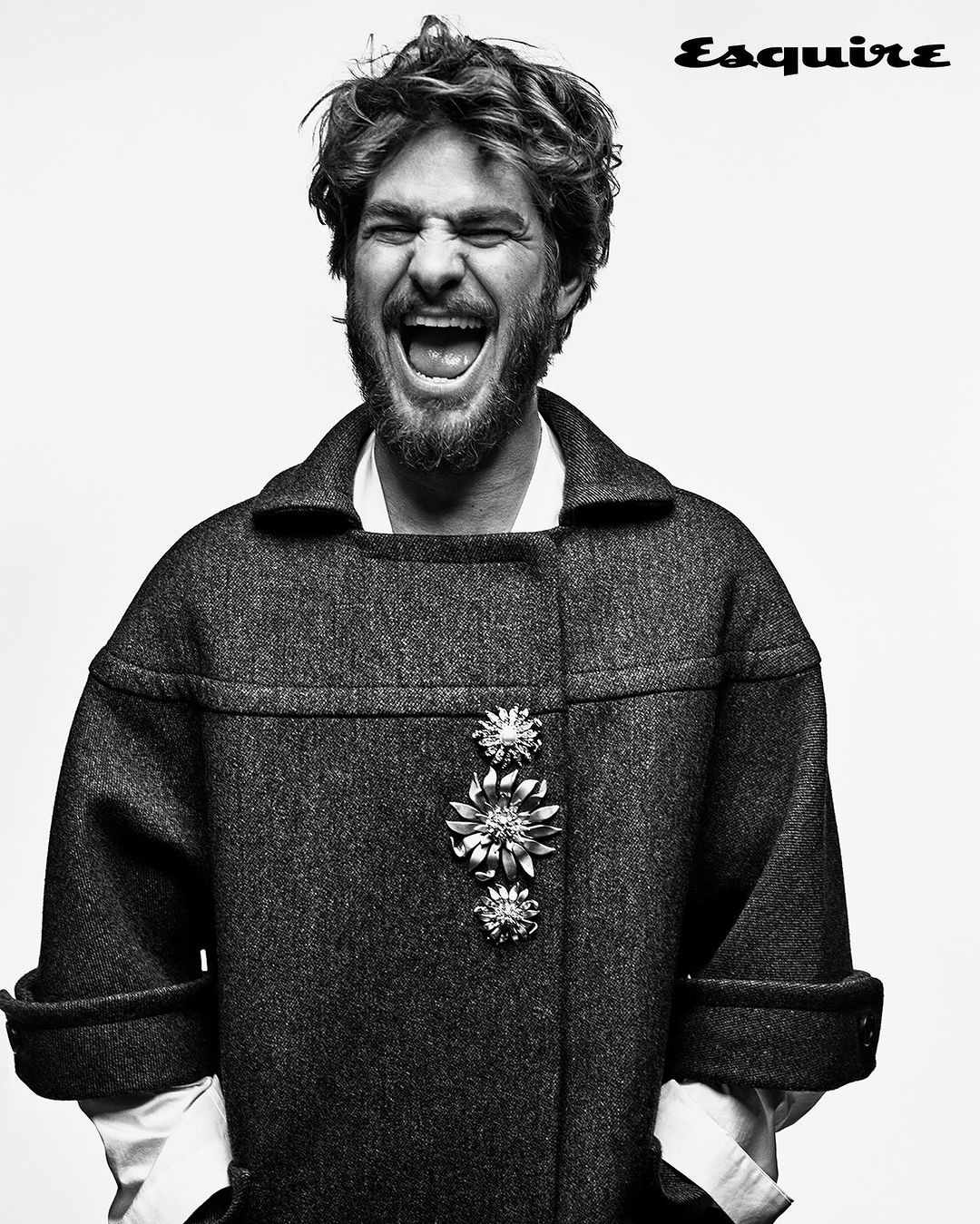 Black and white portrait of a man with curly hair, open mouth, and wearing a coat with brooches on a light background.