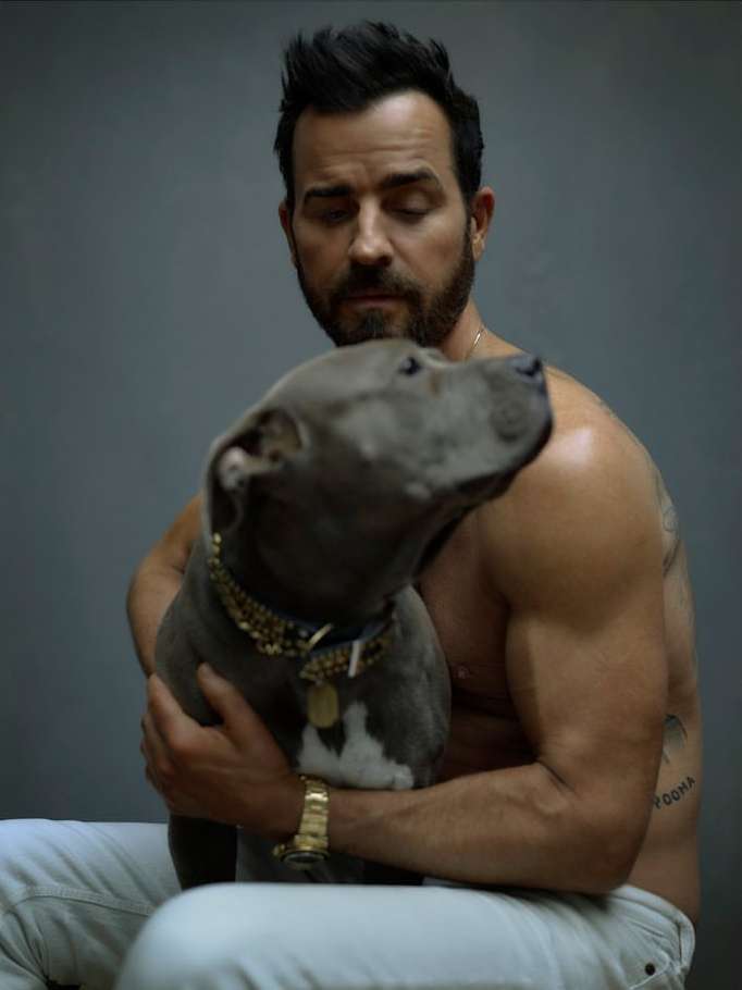 Shirtless man in white jeans sits in a photo studio hugging a pitbull with a gold chain collar, with a neutral gray background.