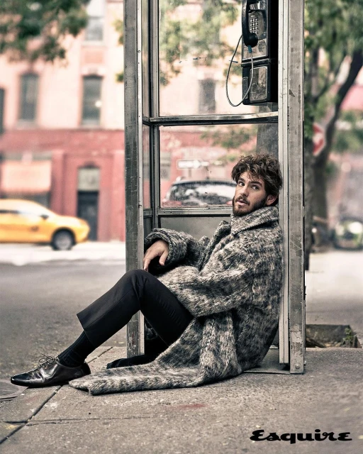 Man in a patterned coat sitting against a phone booth on a city street with buildings and a yellow taxi in the background.