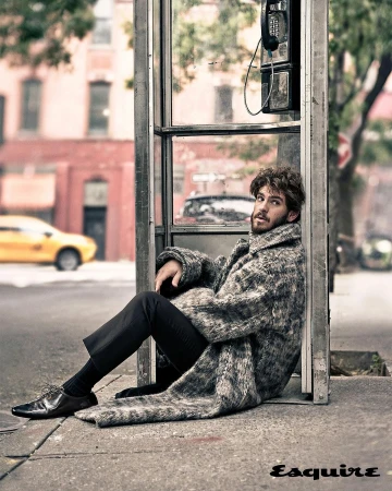 Man in a patterned coat sitting against a phone booth on a city street with buildings and a yellow taxi in the background.