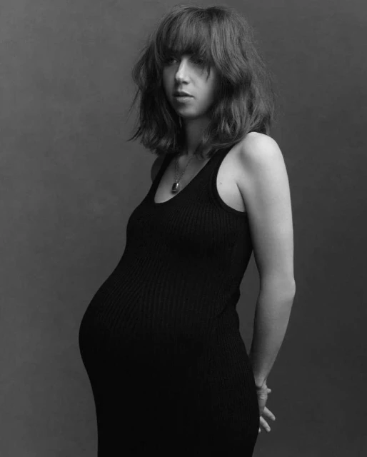 Black and white portrait of a pregnant woman in a black dress standing in front of a solid grey background, with medium shot.