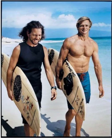 Two men carrying surfboards along a sandy beach near the ocean under a clear sky, photographed by Mark Seliger.