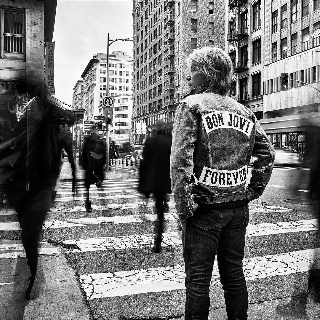 Black and white street photograph of a person wearing a Bon Jovi denim jacket, standing on a crosswalk among blurred pedestrians and city buildings.