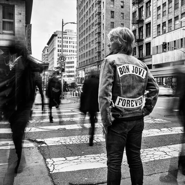Black and white street photograph of a person wearing a Bon Jovi denim jacket, standing on a crosswalk among blurred pedestrians and city buildings.