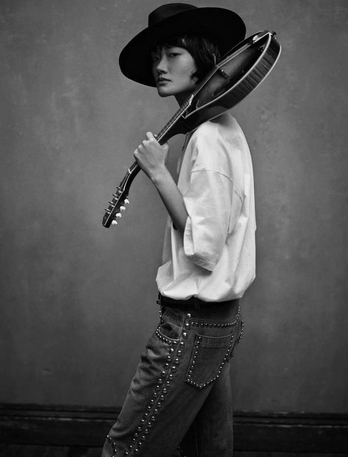 Monochrome portrait of an Asian woman wearing a hat and holding a mandolin, posing against a mottled gray backdrop in a photo studio.