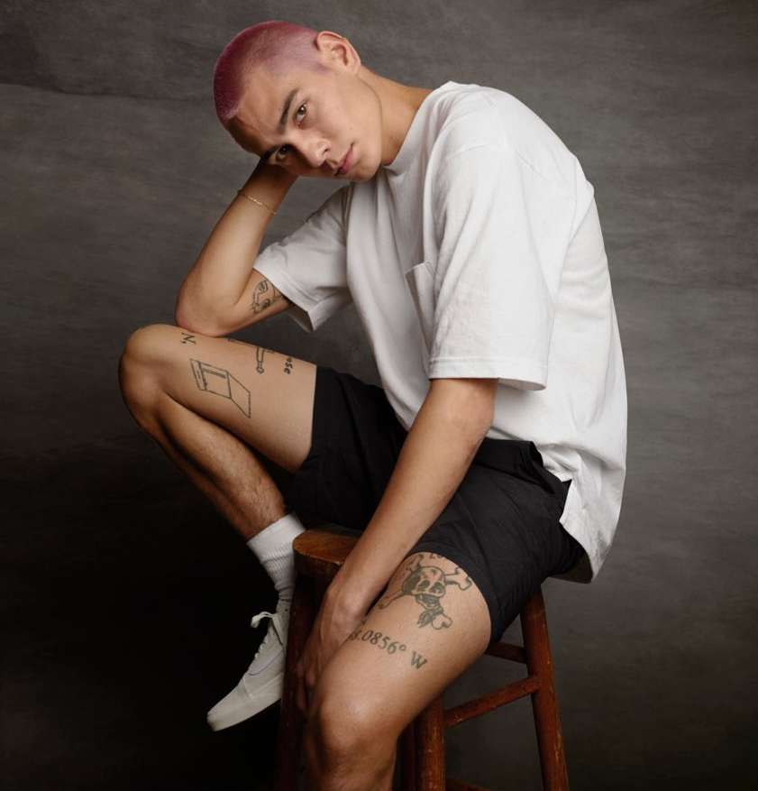 Portrait of a young adult man with a shaved pink head, wearing a white t-shirt and black shorts, sitting on a wooden stool.