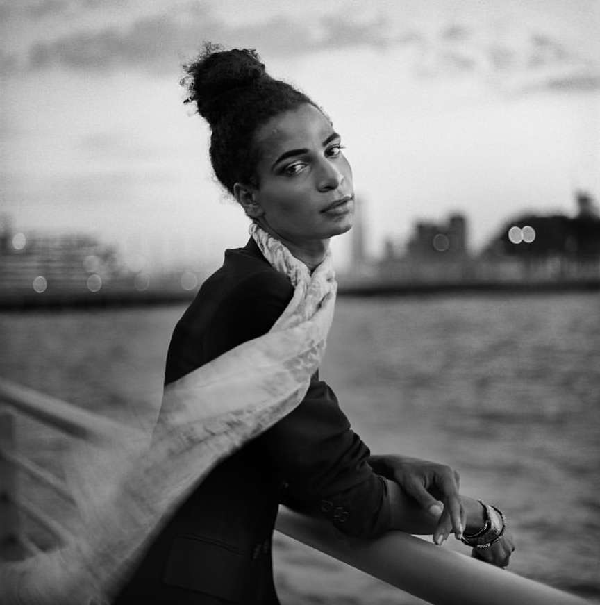 Monochrome portrait of a dark-haired person wearing a dark jacket and white scarf, leaning on a rail with city buildings in background.