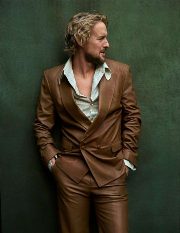 A man in a brown leather suit and patterned shirt poses in front of a dark green wall looking off to the side with a slight smile.