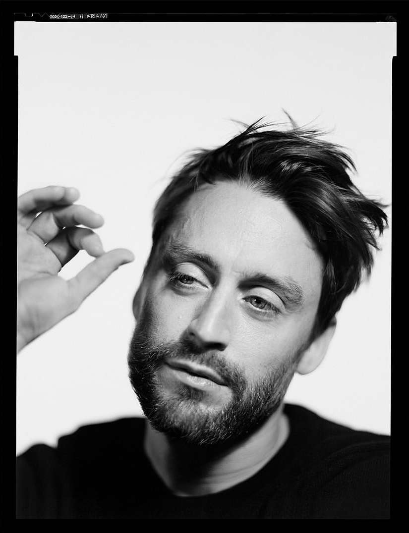 Monochrome portrait of an adult male with a beard, wearing a dark shirt against a light background, with his hand near his face.