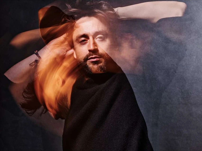 Portrait of a bearded man in a black sweater, with a blurred motion effect on the arms and a dark, smoky background.