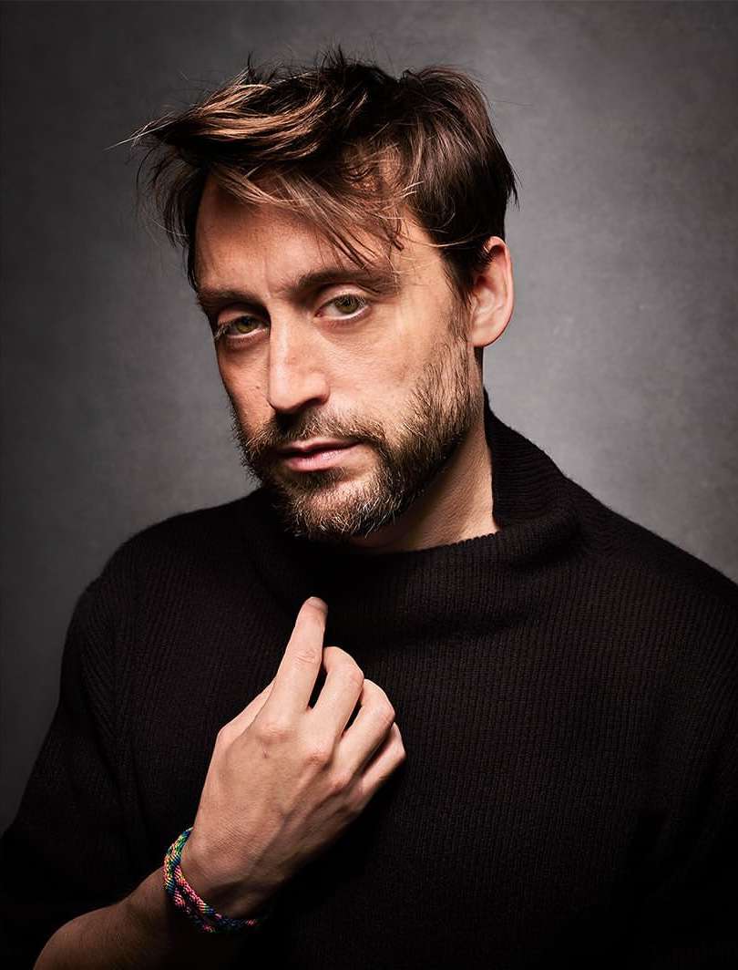 Portrait of a dark-haired adult male wearing a black turtleneck sweater, gazing at the camera in a photo studio setting.
