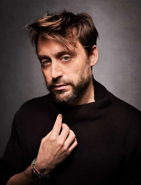 Portrait of a dark-haired adult male wearing a black turtleneck sweater, gazing at the camera in a photo studio setting.