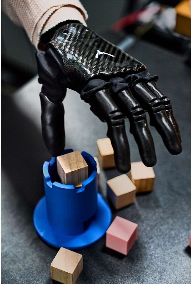 Close-up of a prosthetic hand with carbon fiber detail picking up wooden blocks and placing one in a blue container on a dark surface.