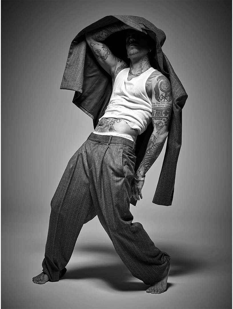 Black and white studio shot of a tatted adult male in wide-legged pants, holding a jacket overhead in a dynamic pose against a grey backdrop.