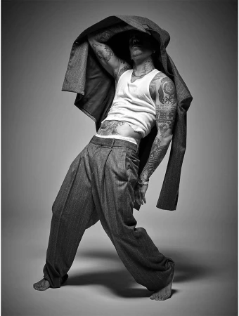 Black and white studio shot of a tatted adult male in wide-legged pants, holding a jacket overhead in a dynamic pose against a grey backdrop.