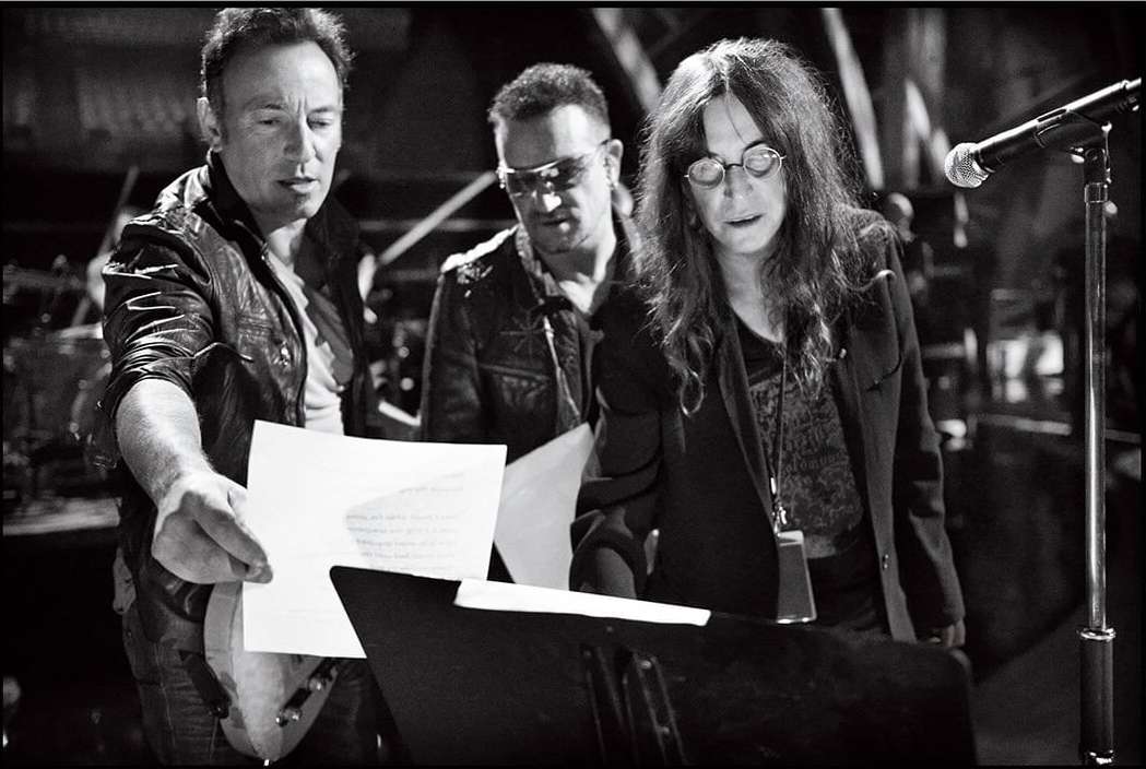 Black and white shot of Bruce Springsteen, Bono, and Patti Smith looking at papers on a music stand, under stage lighting.