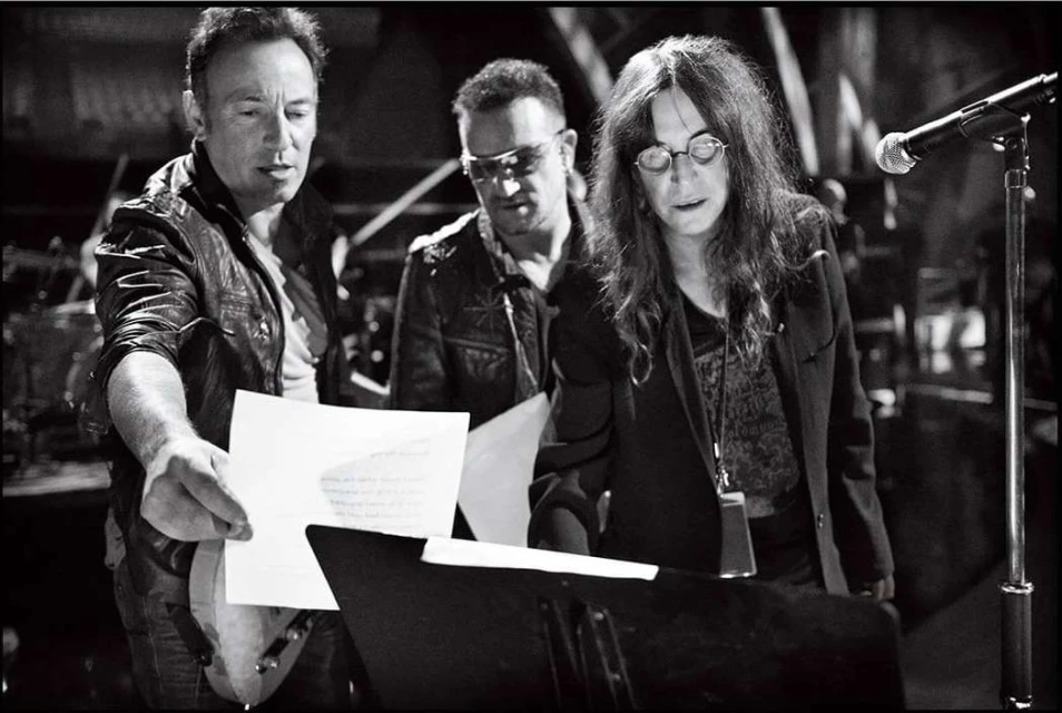 Black and white shot of Bruce Springsteen, Bono, and Patti Smith looking at papers on a music stand, under stage lighting.