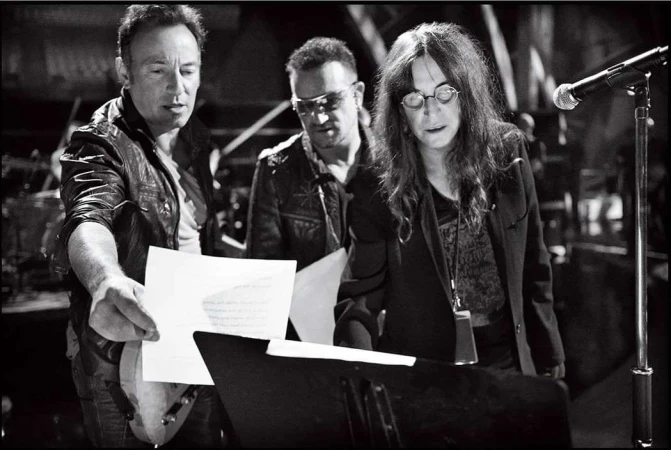 Black and white shot of Bruce Springsteen, Bono, and Patti Smith looking at papers on a music stand, under stage lighting.