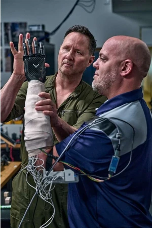 Two adult men examine a robotic prosthetic hand, showcasing technology and healthcare in an indoor lab setting with neutral lighting.