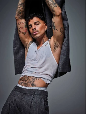Portrait of a tatted man in tank top and striped pants, leaning back with jacket, gray background, fashion, confident pose.