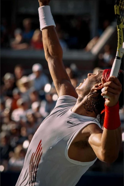A tennis player in a white tank top serves a ball, captured from a low angle, with blurred spectators in the background, showcasing athletic action.