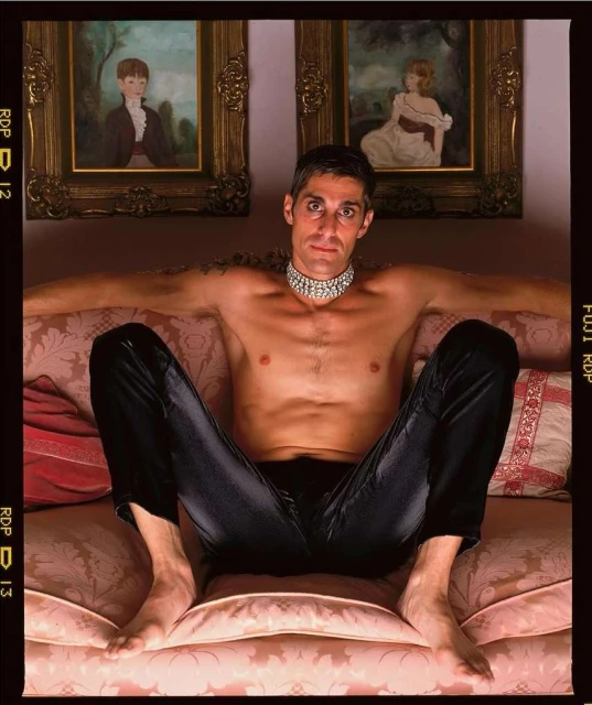 Adult male with black pants sitting on a pink couch with paintings in the background. He's wearing a jeweled choker, looking at the camera.
