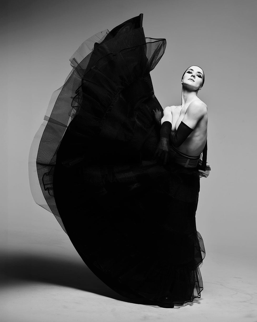 Black and white studio portrait of a female dancer in motion, wearing a dramatic black tulle skirt, posed against a neutral grey backdrop.