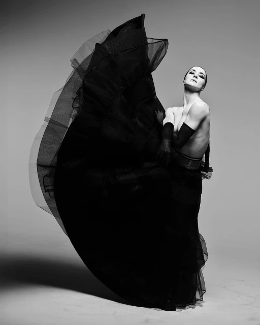 Black and white studio portrait of a female dancer in motion, wearing a dramatic black tulle skirt, posed against a neutral grey backdrop.