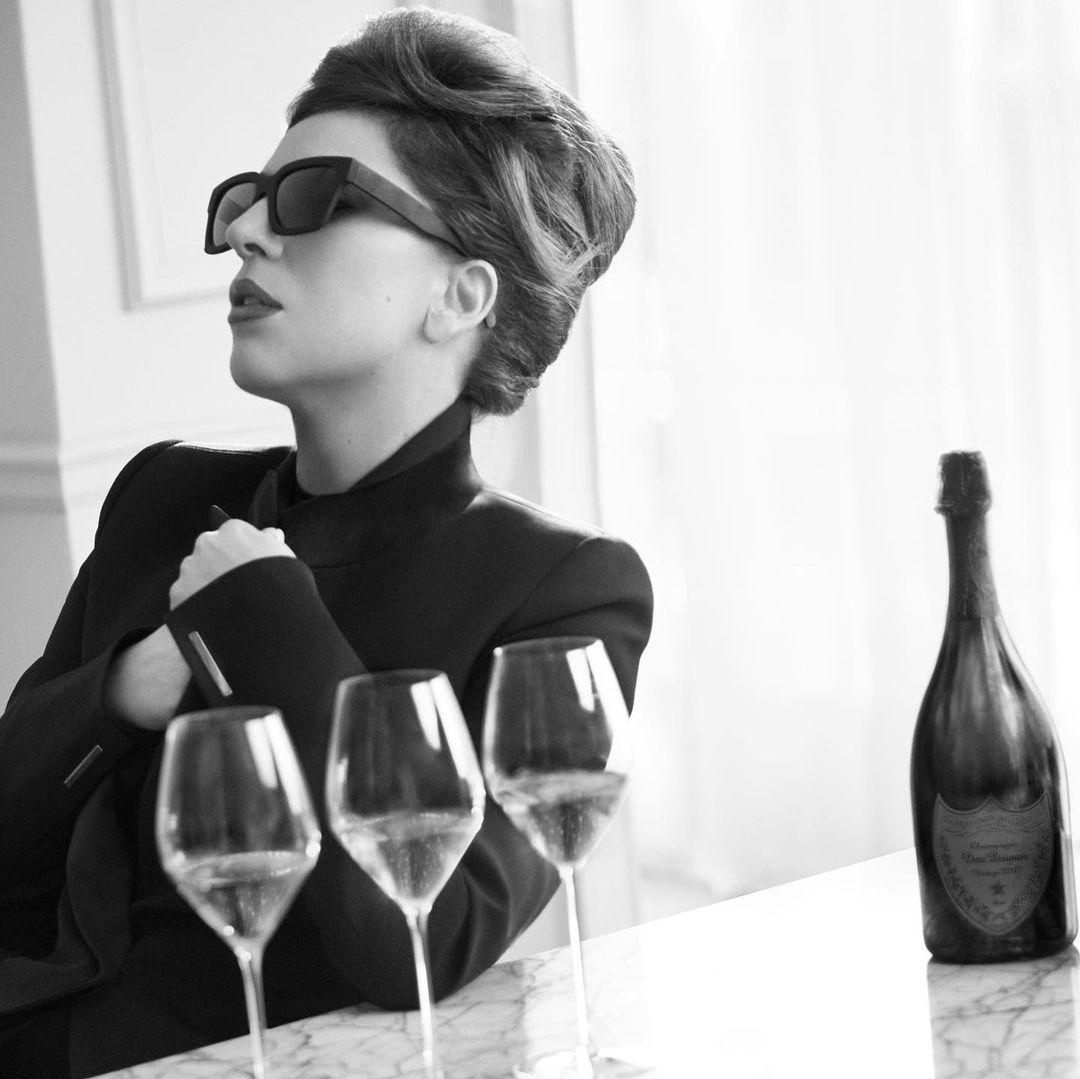 Monochrome portrait of a woman in sunglasses and a blazer at a table with wine glasses and a champagne bottle in a bright indoor setting.