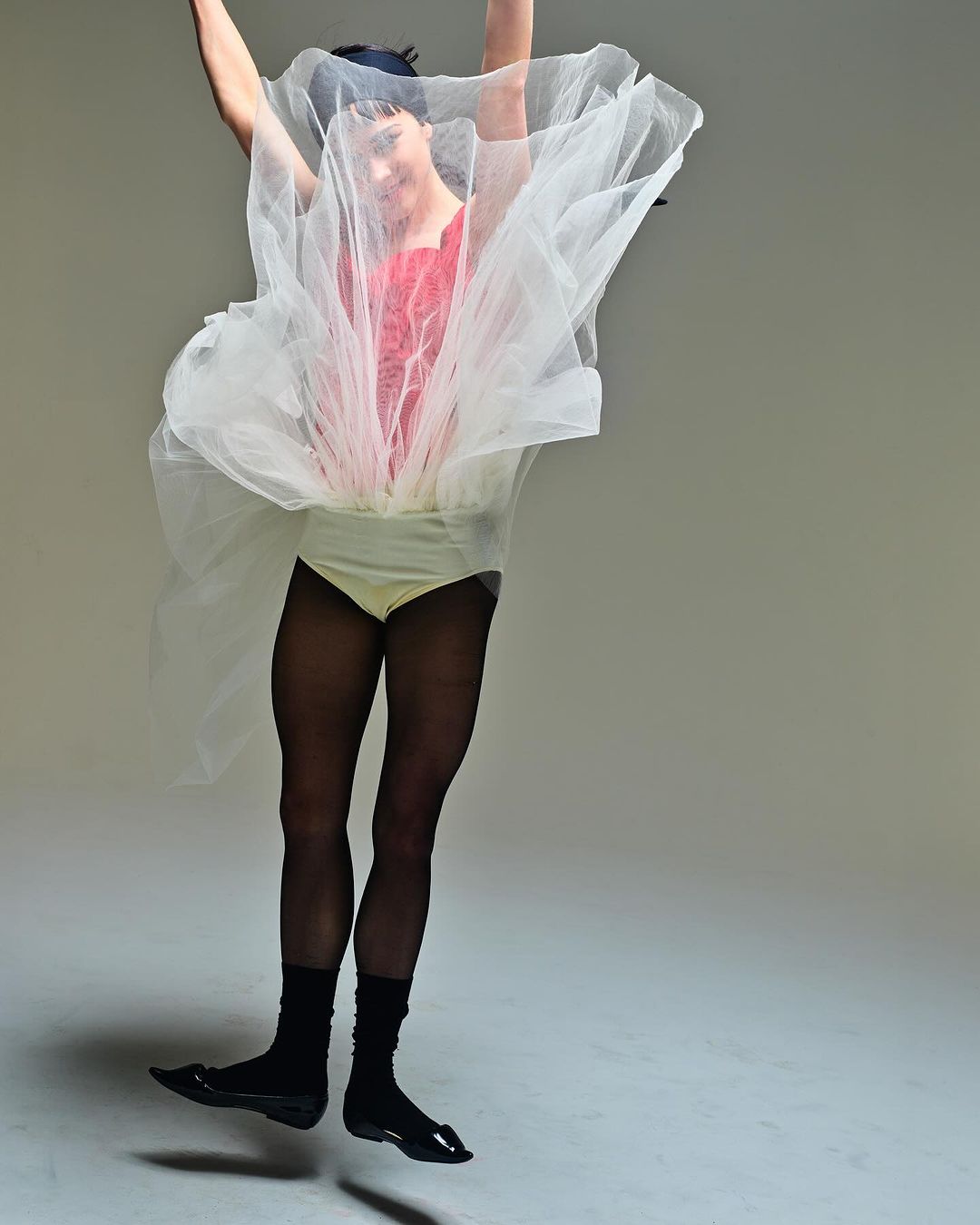 An adult is leaping against a light gray backdrop, wearing black tights, socks, and shiny shoes, a peach leotard, and a red and white tulle skirt.