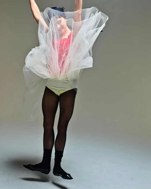 An adult is leaping against a light gray backdrop, wearing black tights, socks, and shiny shoes, a peach leotard, and a red and white tulle skirt.