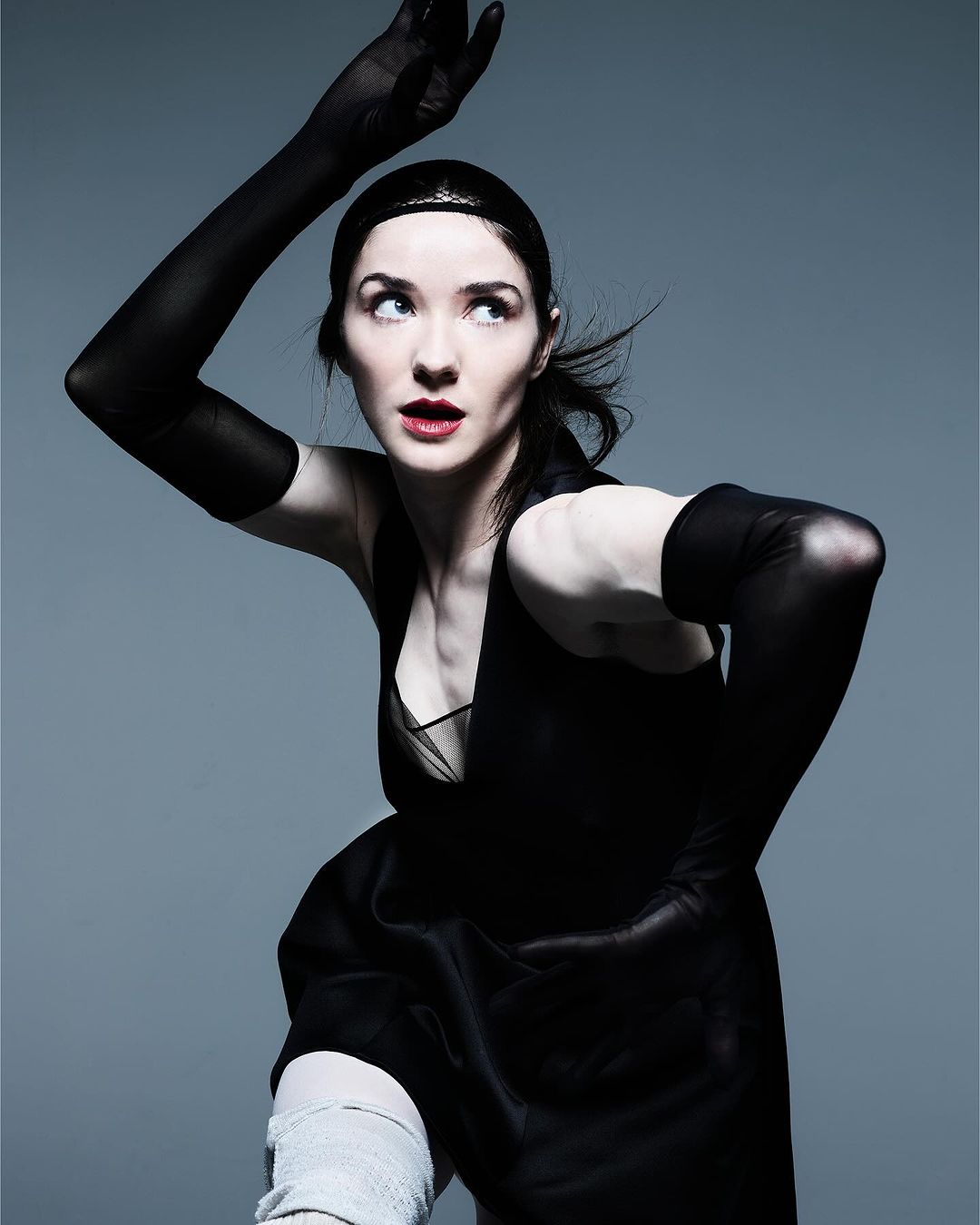 Adult woman posing in a black dress and long black gloves, arm raised, glancing upward against a grey gradient background.