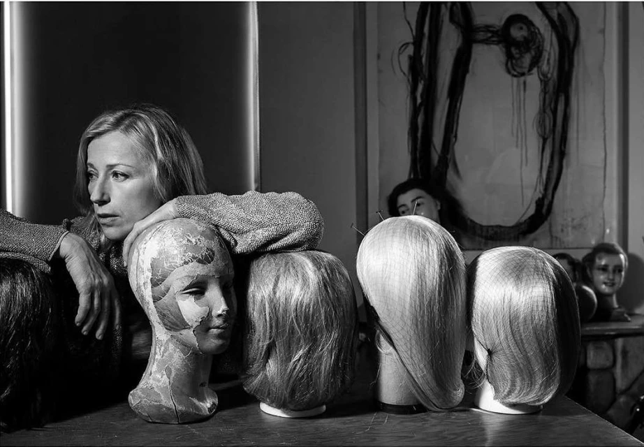 Black and white photo of a blonde woman posing with mannequin heads wearing wigs, set against a textured background with abstract art.