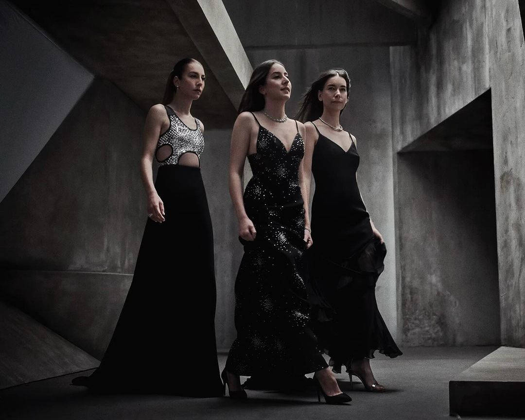 Three women in formal gowns walk in a concrete hallway with geometric architectural features, creating a striking fashion image.