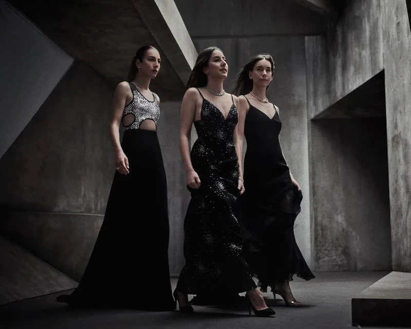 Three women in formal gowns walk in a concrete hallway with geometric architectural features, creating a striking fashion image.