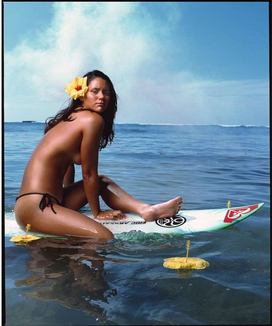 Nude woman with flower in hair sits on surfboard floating in ocean on sunny day with blue sky; visual content for travel, beauty, lifestyle.