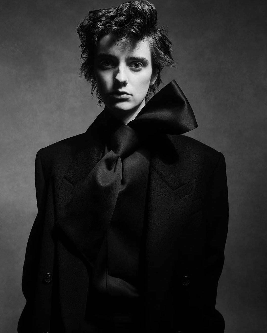 Portrait of a woman with short hair in a black coat and oversized bow tie against a muted grey background in black and white.